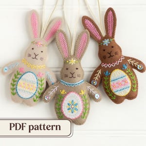 May include: Three felt Easter bunny decorations in beige, brown, and tan, with embroidered details. The bunnies have pink-lined ears, floral accents, and an embroidered egg design on their bellies. The text "PDF pattern" is at the bottom.