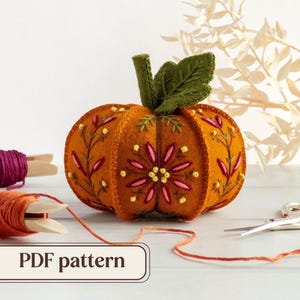 May include: An orange felt pumpkin with embroidered floral details, a green stem, and leaf. The pumpkin is surrounded by spools of thread in shades of orange and purple, scissors, and the text "PDF pattern".