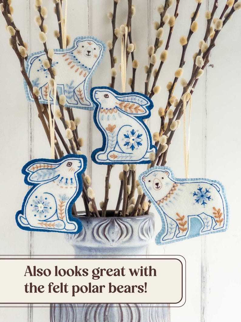 Two embroidered felt winter hares and two polar bears are hanging in branches of pussy willow in a grey vase. They are all made of blue and white wool felt, with hand-embroidered details in a folk-art style. Overlaid text at the bottom of the image reads: "Also looks great with the felt polar bears!"