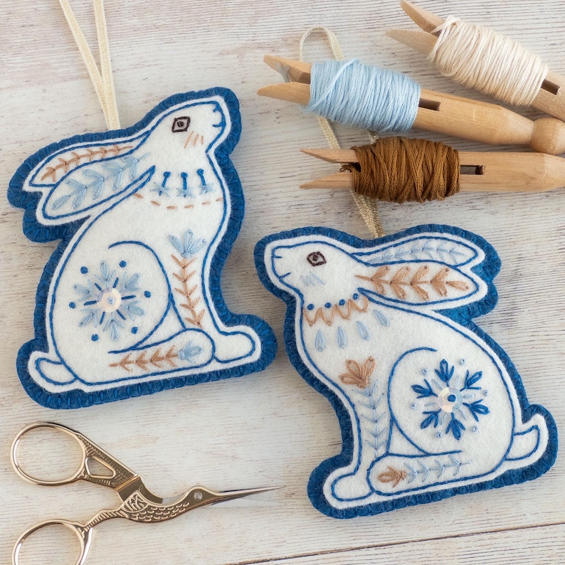 Two blue and felt winter hare ornaments with embroidered details are arranged on a white-washed wooden surface. They have hangers made of gold ribbon and snowflake motifs on their back legs. Embroidery thread and stork scissors are also in shot.