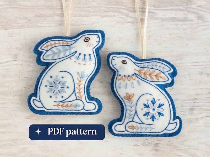 Two embroidered felt winter hare ornaments are arranged on a white-washed wooden surface. They are made of blue and white wool felt, with hand-embroidered details in a folk-art style. Overlaid text reads "PDF pattern".