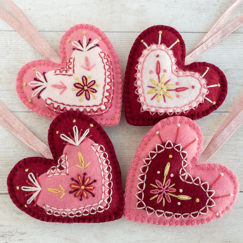 Four hand-embroidered wool felt heart ornaments are arranged on a white-washed wooden surface. They are made of pink and red felt and they have sparkly pink ribbon hangers.
