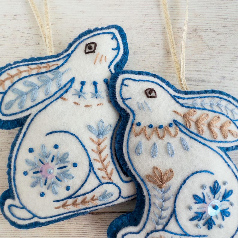 Close-up photo of two handmade wool felt hare ornaments, showing hand-embroidered details in blue and light brown. They have snowflake motifs on their hind legs.