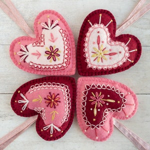 Four wool felt heart ornaments with hand embroidered details in a folk art style. Each heart has a different embroidered design with pink, red, and gold thread.