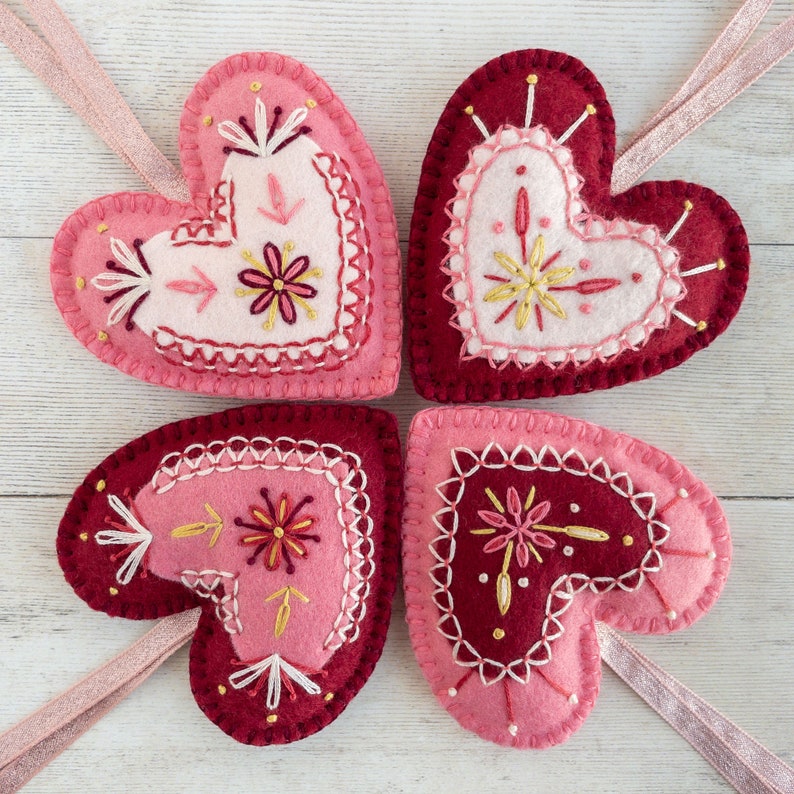 Four wool felt heart ornaments with hand embroidered details in a folk art style. Each heart has a different embroidered design with pink, red, and gold thread.