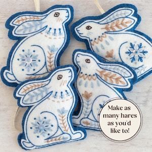 Four blue and white felt hare ornaments are arranged on a white-washed wooden surface. They have hand-stitched details in a folk art style. Overlaid text reads "Make as many hares as you'd like to!".