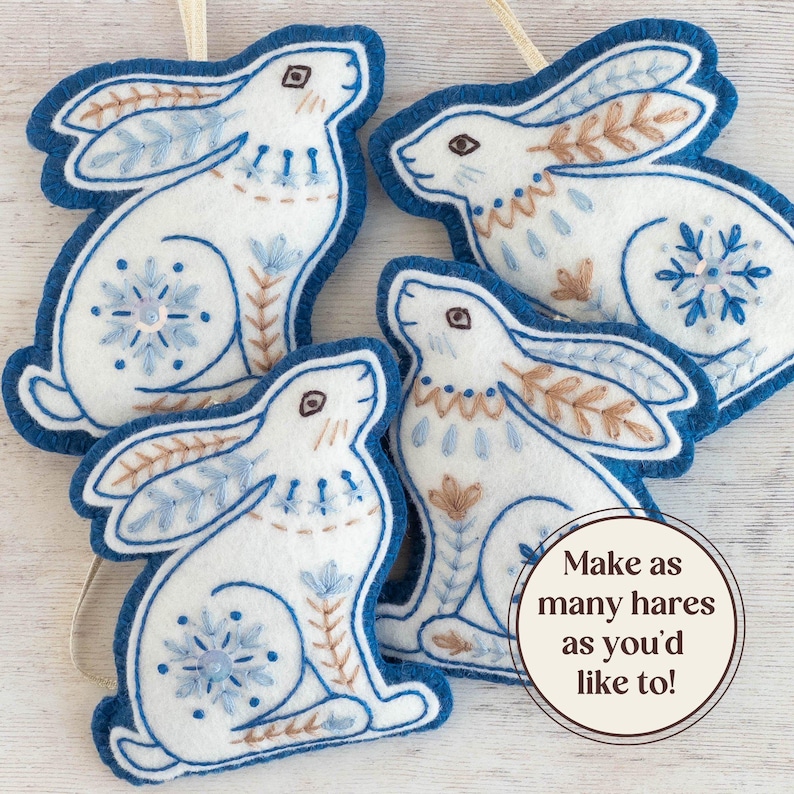 Four blue and white felt hare ornaments are arranged on a white-washed wooden surface. They have hand-stitched details in a folk art style. Overlaid text reads "Make as many hares as you'd like to!".