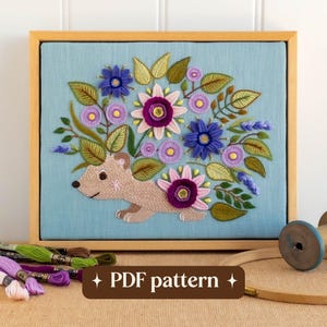 May include: Embroidered artwork featuring a light brown animal with a floral design. The embroidery is set in a wooden frame against a light blue background. The flowers are purple, pink, and blue. The text "PDF pattern" is at the bottom.