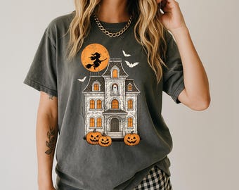 May include: Dark gray t-shirt featuring a Halloween design. The graphic includes a haunted house, pumpkins, bats, and a witch flying in front of a full moon. The shirt is a relaxed fit.