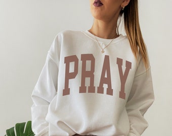 Puede incluir: Sudadera blanca con un estampado marrón de "PRAY" en la parte delantera. La sudadera se lleva con pantalones de chándal marrones.
