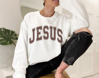 May include: A white long-sleeve sweatshirt with the word "JESUS" printed in brown block letters across the chest. The sweatshirt is paired with black distressed jeans. The overall style is casual and comfortable.