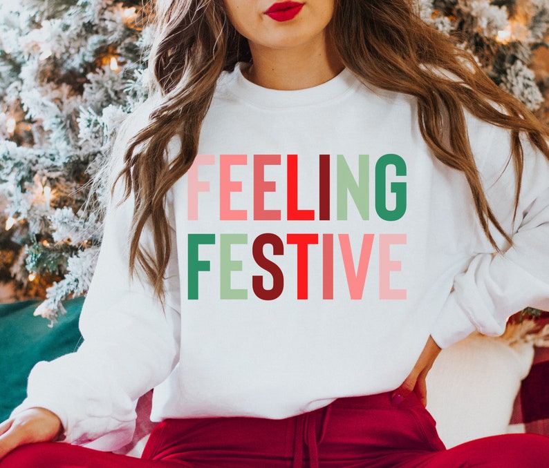 May include: White sweatshirt with the text "FEELING FESTIVE" in colorful letters. The letters are in shades of red, green, pink, and white.