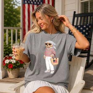 May include: A gray t-shirt featuring a cartoon bear wearing sunglasses, a red and white striped shirt, white pants, and carrying a straw tote bag with an American flag. The bear is wearing a white bandana. The shirt is a relaxed fit.
