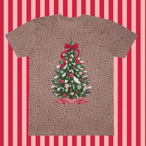 May include: A brown t-shirt with a leopard print design. The shirt features a Christmas tree design with red, pink, and green baubles, a red bow, and a red ribbon. The background is a red and pink striped pattern.