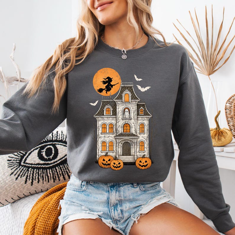 May include: Dark gray sweatshirt featuring a Halloween-themed graphic. The design includes a haunted house, a witch on a broomstick in front of a full moon, bats, and jack-o'-lanterns. The sweatshirt is a casual, long-sleeved garment.