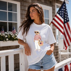 May include: White t-shirt featuring a bear wearing sunglasses, a red and white striped shirt, white pants, and carrying a straw tote bag with an American flag. The bear is wearing a polka dot scarf. The background includes an American flag.