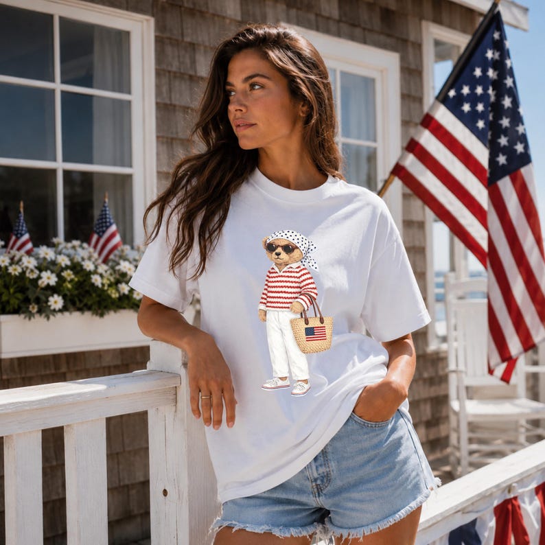 May include: White t-shirt featuring a bear wearing sunglasses, a red and white striped shirt, white pants, and carrying a straw tote bag with an American flag. The bear is wearing a polka dot scarf. The background includes an American flag.