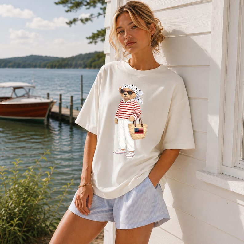 May include: Cream-colored t-shirt featuring a cartoon bear wearing sunglasses, a red and white striped shirt, white pants, and a polka-dot scarf. The bear is carrying a straw tote bag with an American flag design. The model is wearing blue and white striped shorts.