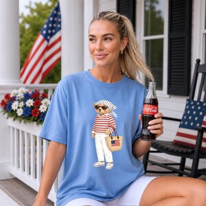 May include: Light blue t-shirt featuring a bear wearing sunglasses, a red and white striped shirt, white pants, and a straw bag with an American flag. The bear is holding the bag and wearing a white and black polka dot scarf. A Coca-Cola bottle is held in the hand.