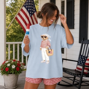 May include: Light blue t-shirt featuring a bear wearing sunglasses, a red and white striped shirt, white pants, and a straw bag with an American flag. The bear is wearing a white bandana with black polka dots. The shirt is paired with red and white checkered shorts.