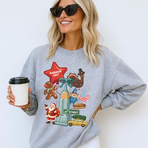May include: A light gray sweatshirt featuring a colorful graphic of a star-shaped balloon with "Parade Day" text, a gingerbread man, Santa Claus, a turkey, an elf, the Empire State Building, a yellow taxi, and an American flag.