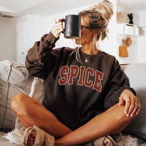 May include: A woman wearing a brown sweatshirt with the word "SPICE" printed in red on the front. She is sitting on a couch and holding a black mug.