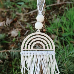 May include: A wooden rainbow ornament with a macrame fringe hanging from a branch. The ornament is painted white and has a natural wood finish.