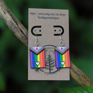 May include: A pair of rainbow pride flag earrings with a white heart on each. The earrings are made of wood and have silver hooks. The earrings are displayed on a brown card with the text "Knottyworkdesigns" and a website address.