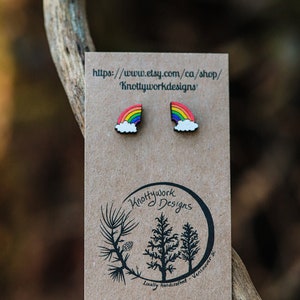 Pu&ograve; includere: Un paio di orecchini in legno a forma di arcobaleno e nuvola su una carta marrone con il testo "Knottywork Designs" e "Artigianato locale - Vancouver, BC".