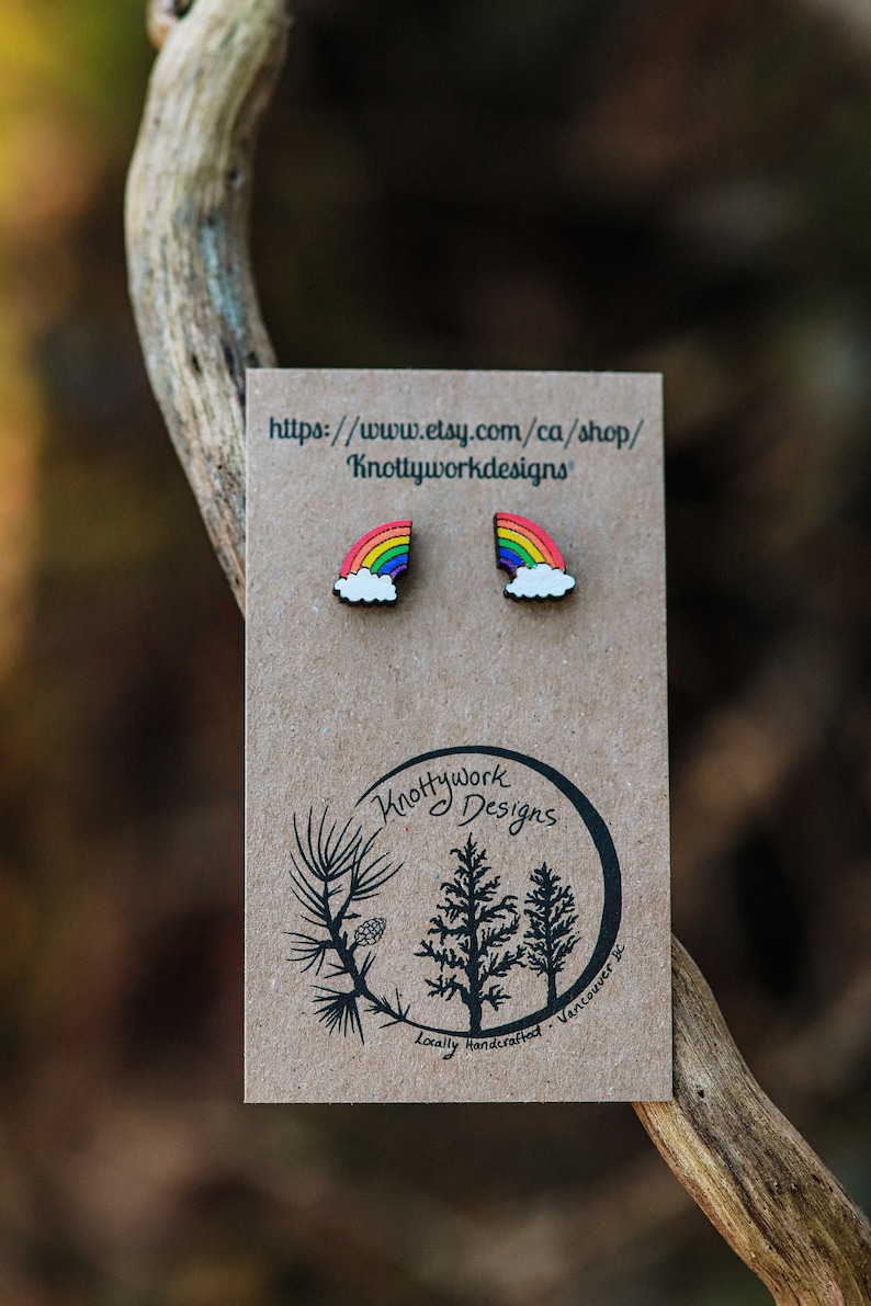 Pu&ograve; includere: Un paio di orecchini in legno a forma di arcobaleno e nuvola su una carta marrone con il testo "Knottywork Designs" e "Artigianato locale - Vancouver, BC".