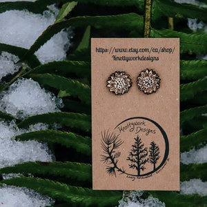 May include: A pair of wooden earrings featuring a white sunflower design. The earrings are displayed on a brown card with the text "Knottywork Designs" and a website address: "https://www.etsy.com/ca/shop/KnottyworkDesigns".