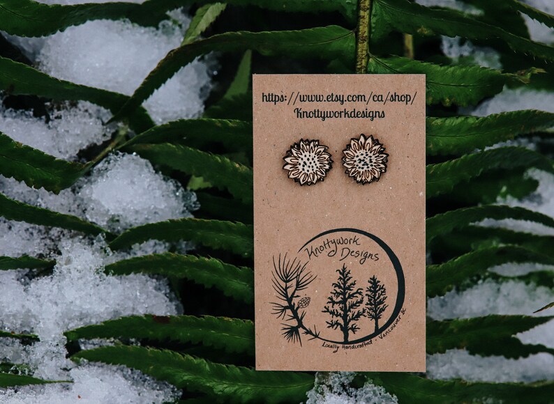 May include: A pair of wooden earrings featuring a white sunflower design. The earrings are displayed on a brown card with the text "Knottywork Designs" and a website address: "https://www.etsy.com/ca/shop/KnottyworkDesigns".