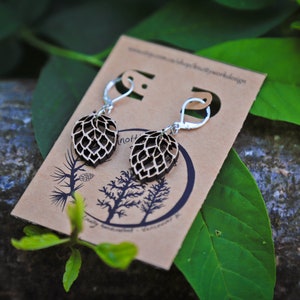 May include: A pair of wooden earrings with a leaf design. The earrings are brown and have silver hooks. The earrings are on a brown card with the text "Knotty Butterfly Woodwork Design" printed on it.
