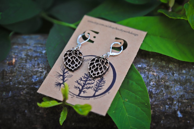 May include: A pair of wooden earrings with a leaf design. The earrings are brown and have silver hooks. The earrings are on a brown card with the text "Knotty Butterfly Woodwork Design" printed on it.