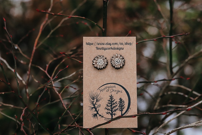 May include: A pair of wooden sunflower earrings on a brown card with the text "https://www.etsy.com/ca/shop/Knottyworkdesigns" and "Knottywork Designs".