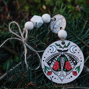 Może przedstawiać: Drewniana ozdoba z ręcznie malowanym wzorem ćmy z czerwonymi różami i półksiężycami. Ozdoba jest nawleczona na sznurek z drewnianymi koralikami.