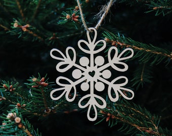 Op de afbeelding: Een witte houten sneeuwvlok ornament met een klein hart in het midden, hangend aan een bruin touwtje aan een groene dennentak.