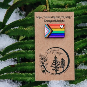 May include: A wooden pin featuring a rainbow pride flag with a white heart in the top right corner. The pin is on a brown card with the text "Knottywork Designs" and a website address: "https://www.etsy.com/ca/shop/KnottyworkDesigns".