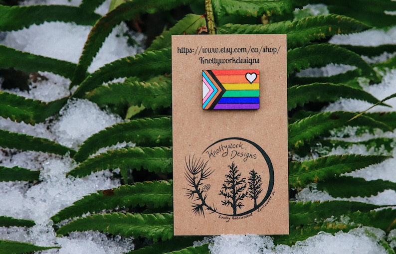 May include: A wooden pin featuring a rainbow pride flag with a white heart in the top right corner. The pin is on a brown card with the text "Knottywork Designs" and a website address: "https://www.etsy.com/ca/shop/KnottyworkDesigns".