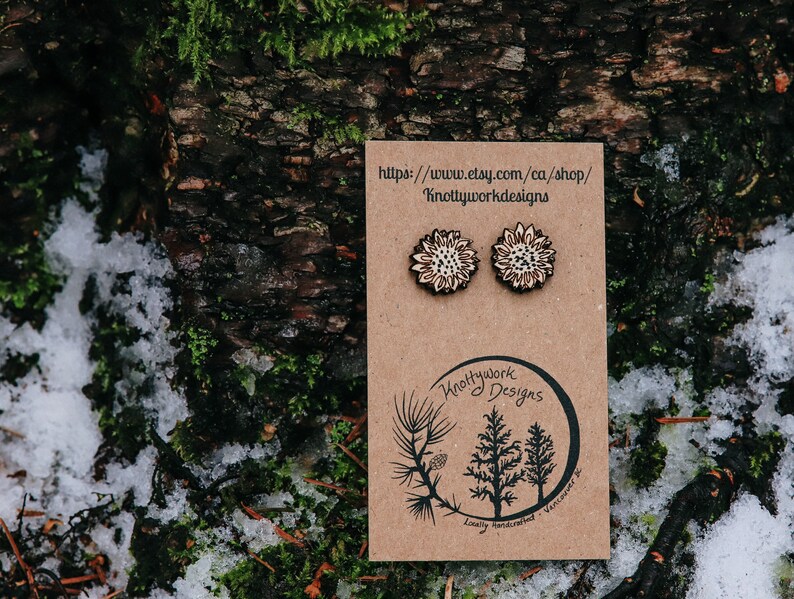 May include: A pair of wooden earrings with a sunflower design. The earrings are on a brown card with the text "Knottywork Designs" and a website address.