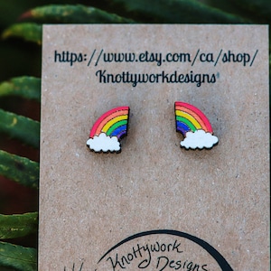Pu&ograve; includere: Un paio di orecchini in legno arcobaleno con un design a nuvola bianca. Gli orecchini sono esposti su una carta marrone con il testo "https://www.etsy.com/ca/shop/Knottyworkdesigns".