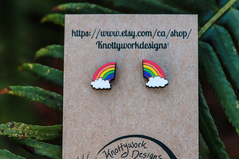 Pu&ograve; includere: Un paio di orecchini in legno arcobaleno con un design a nuvola bianca. Gli orecchini sono esposti su una carta marrone con il testo "https://www.etsy.com/ca/shop/Knottyworkdesigns".