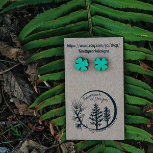 Op de afbeelding: Een paar groene vierbladige klaver-oorbellen op een bruine kaart met de tekst "https://www.etsy.com/ca/shop/KnottyworkDesigns" en "Knottywork Designs" in een cirkel.