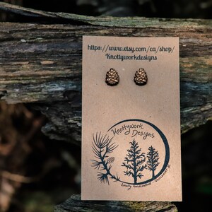 K&ouml;nnte beinhalten: Ein Paar Ohrstecker aus Holz in Form von Tannenzapfen auf einer braunen Karte mit dem Text "Knottywork Designs" und einer Website-Adresse. Die Ohrstecker sind hellbraun und haben eine nat&uuml;rliche Holzoptik.