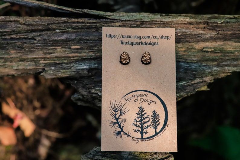 K&ouml;nnte beinhalten: Ein Paar Ohrstecker aus Holz in Form von Tannenzapfen auf einer braunen Karte mit dem Text "Knottywork Designs" und einer Website-Adresse. Die Ohrstecker sind hellbraun und haben eine nat&uuml;rliche Holzoptik.
