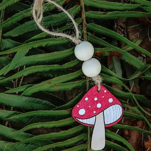 May include: A red and white mushroom-shaped wooden ornament with a white string and two white wooden beads. The ornament is lying on a green fern leaf.