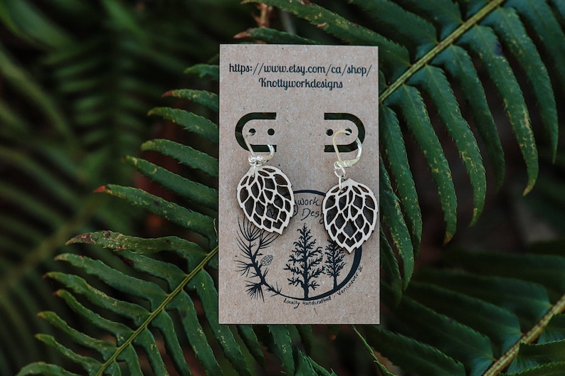 May include: A pair of wooden earrings shaped like a hop leaf. The earrings are on a brown card with the text "https://www.etsy.com/ca/shop/Knottyworkdesigns" printed on it.