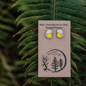 May include: A pair of wooden earrings shaped like beer mugs with a yellow beer and white foam. The earrings are on a brown card with the text "Knottywork Designs" and a website address.