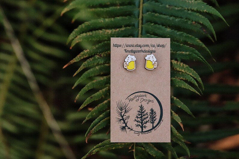 May include: A pair of wooden earrings shaped like beer mugs with a yellow beer and white foam. The earrings are on a brown card with the text "Knottywork Designs" and a website address.