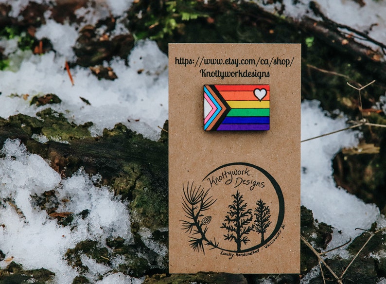 May include: A wooden pin featuring the Progress Pride flag with a white heart in the top right corner. The pin is on a brown card with the text "Knottywork Designs" and a website address.